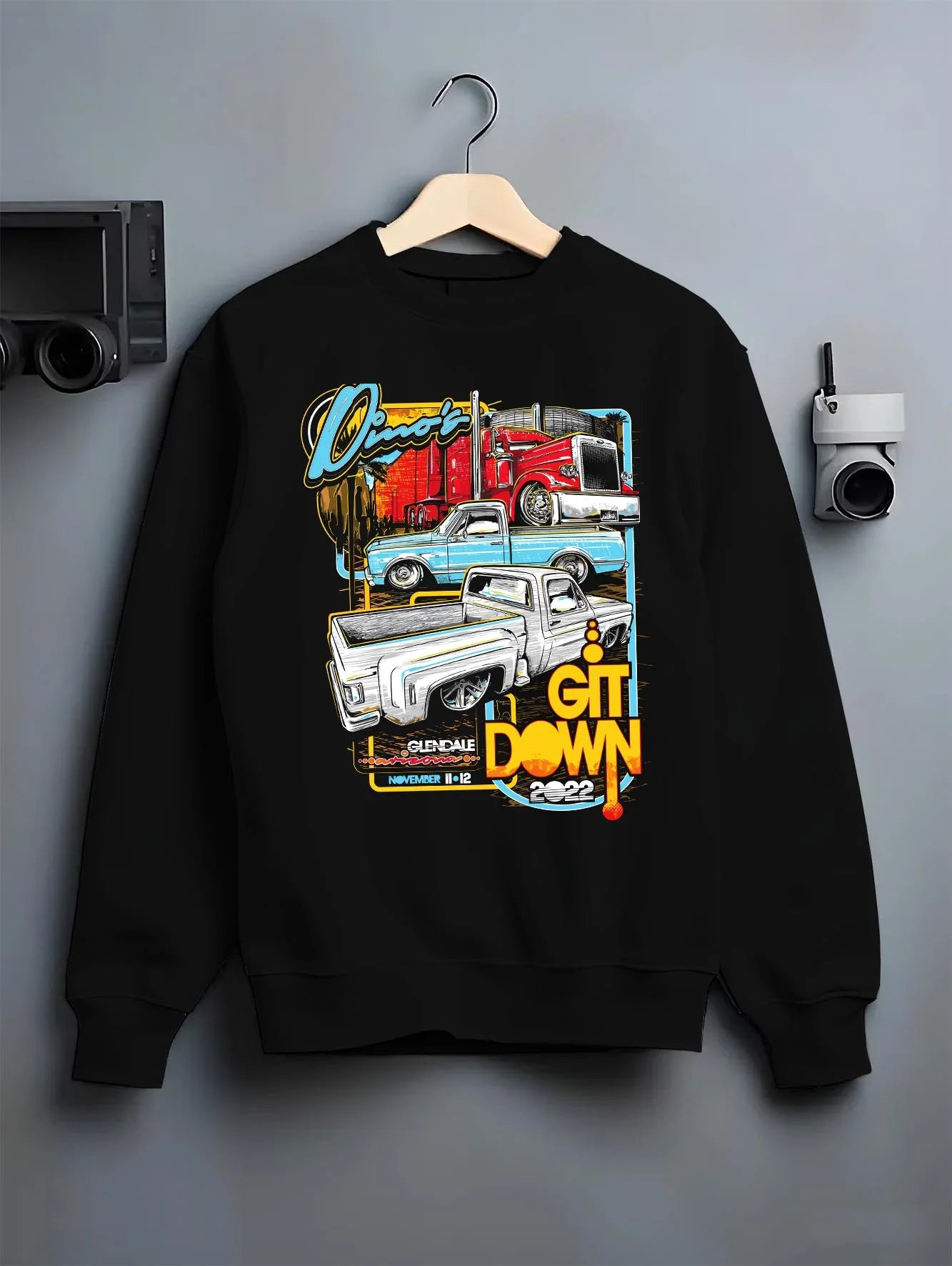 Black crewneck sweatshirt on a hanger against a grey wall, featuring street retro classic show racing performance graphic.