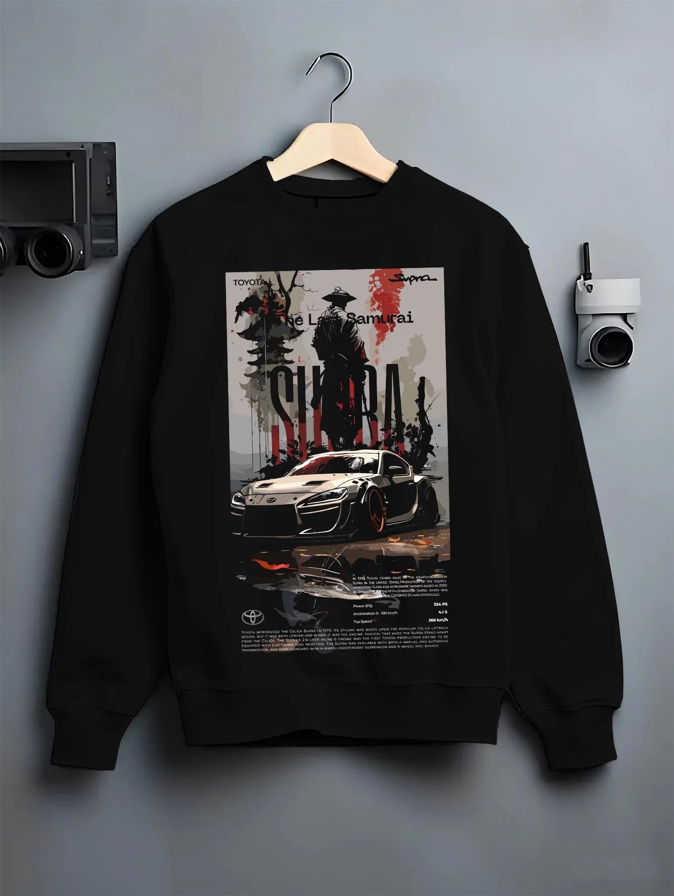 Black crewneck sweatshirt on a hanger against a grey wall, featuring Toyota Supra racing jdm japanese tuning street graphic.