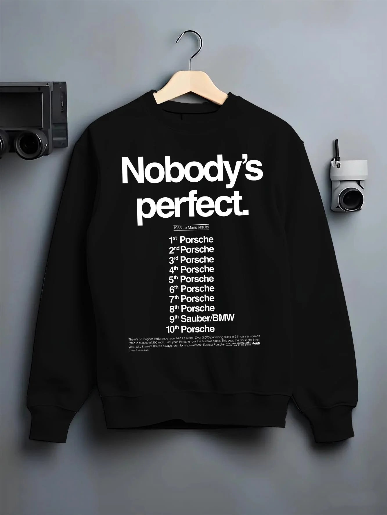Black crewneck sweatshirt on a hanger against a grey wall, featuring Porsche rs racing le mans motorsport street graphic.